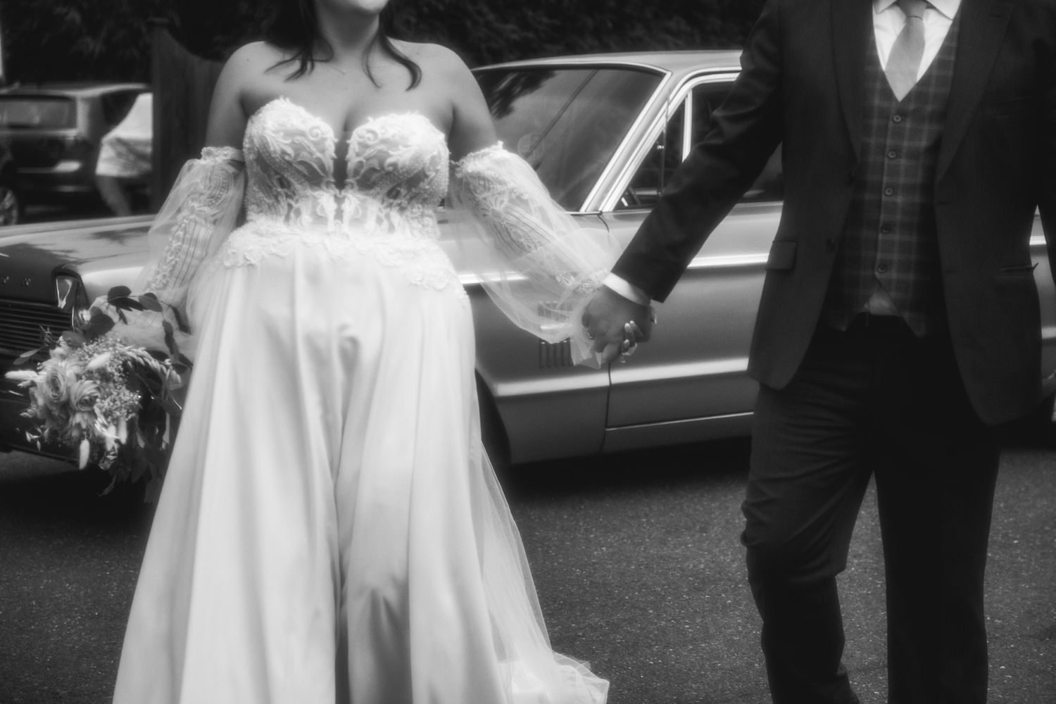 Candid black and white wedding portrait showing a couple holding hands outdoors near classic cars.