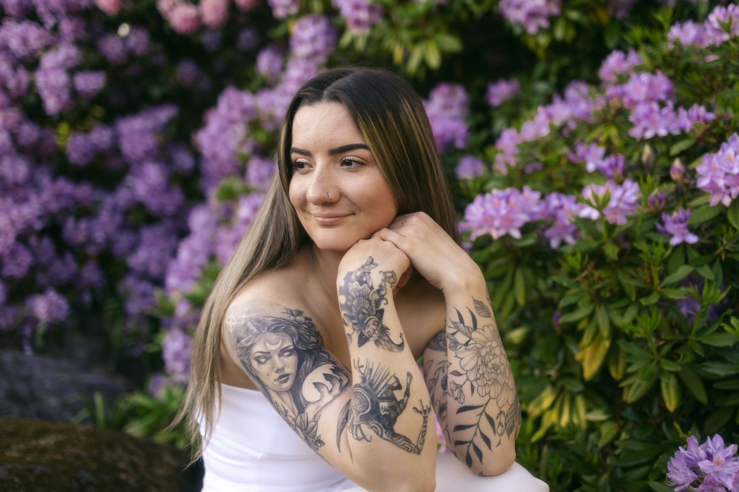Someone in a white top poses in front of blooming purple rhododendrons showing their artistic arm tattoos.