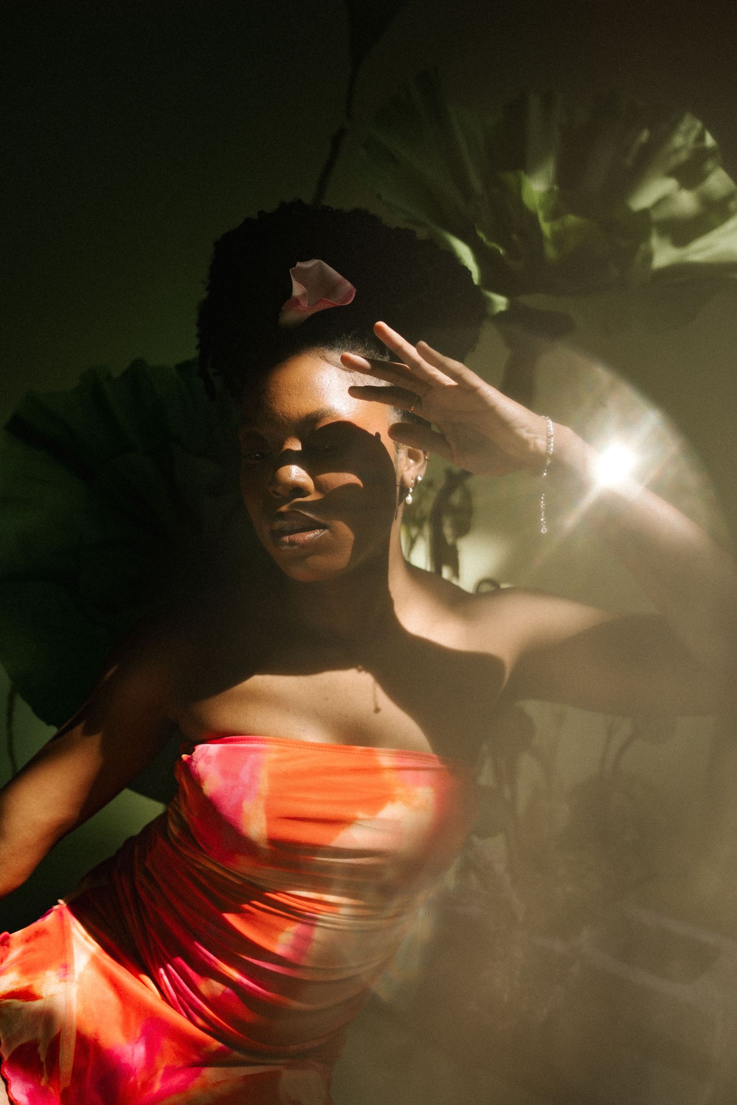 A Black woman in a red strapless dress poses dramatically with raised hand against a dark, moody background with tropical leaves.