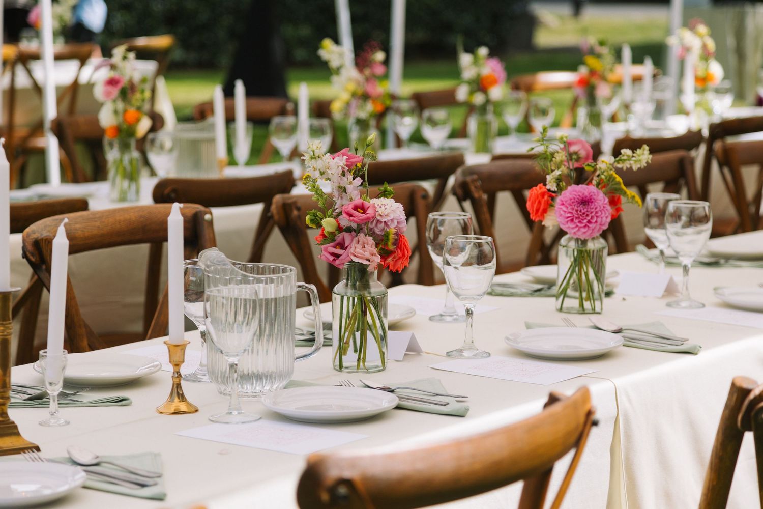 Elegant outdoor wedding reception table settings with wooden chairs, white linens and delicate floral centerpieces.