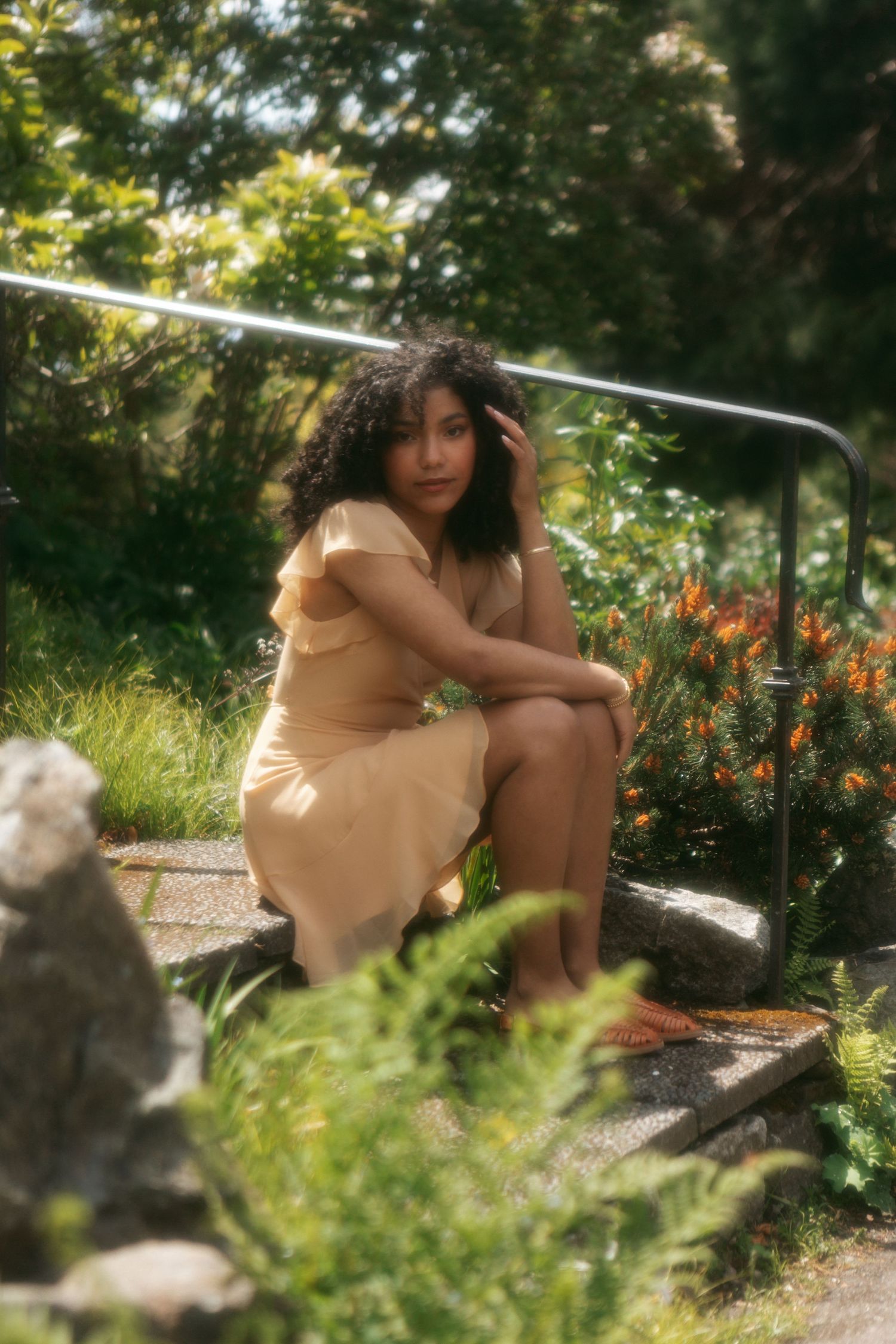 A person in a tan dress sits on outdoor steps surrounded by lush garden greenery and orange flowers on a sunny day.