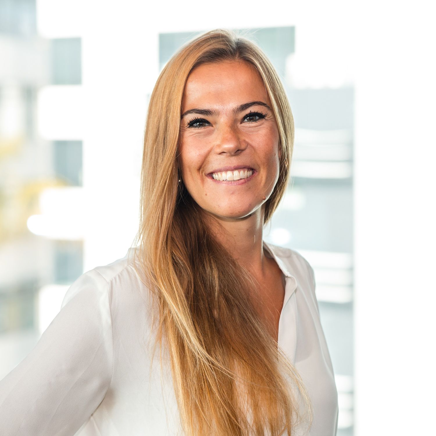 Professional headshot of a person in white blouse against a bright office background.