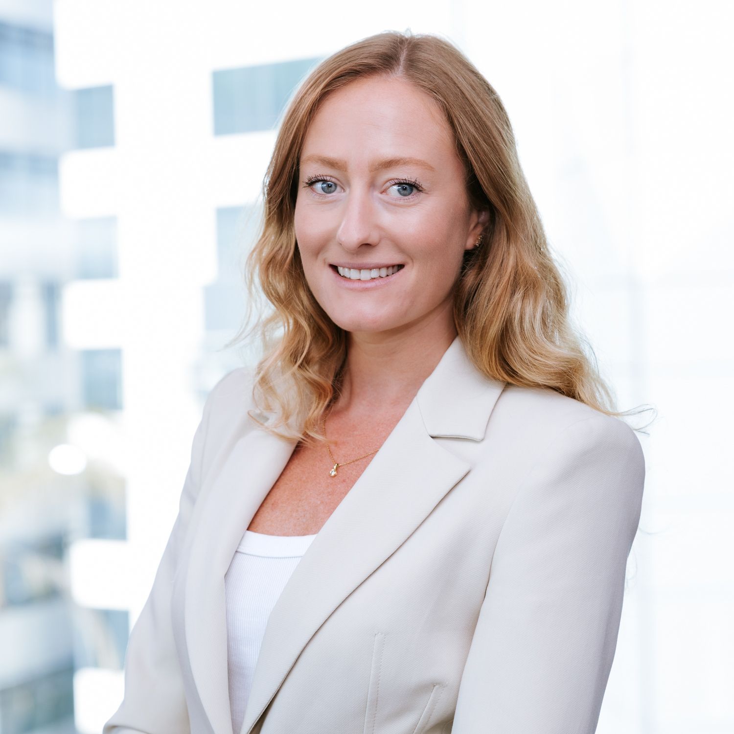 Professional headshot of someone in a white blazer and blouse against a bright office background.