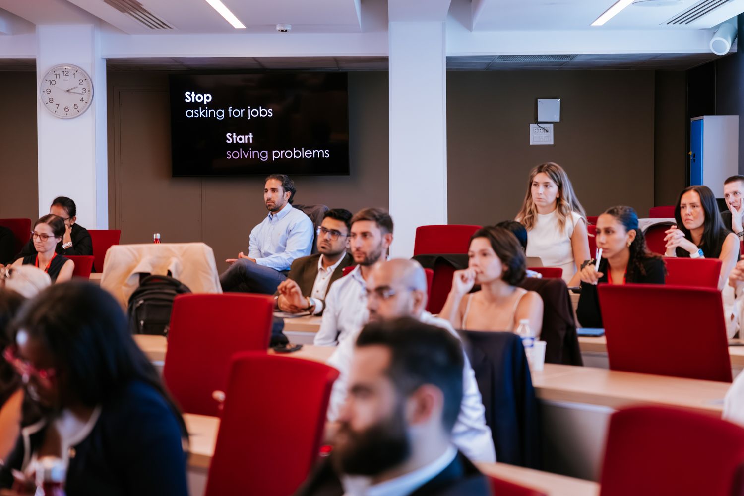 A panoramic view of a modern conference room with red chairs and people seated during a presentation.