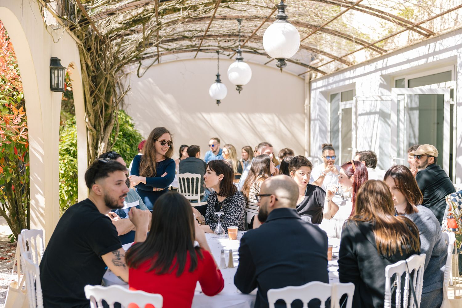 People gathered for a social event under a covered patio with hanging lights and vines.