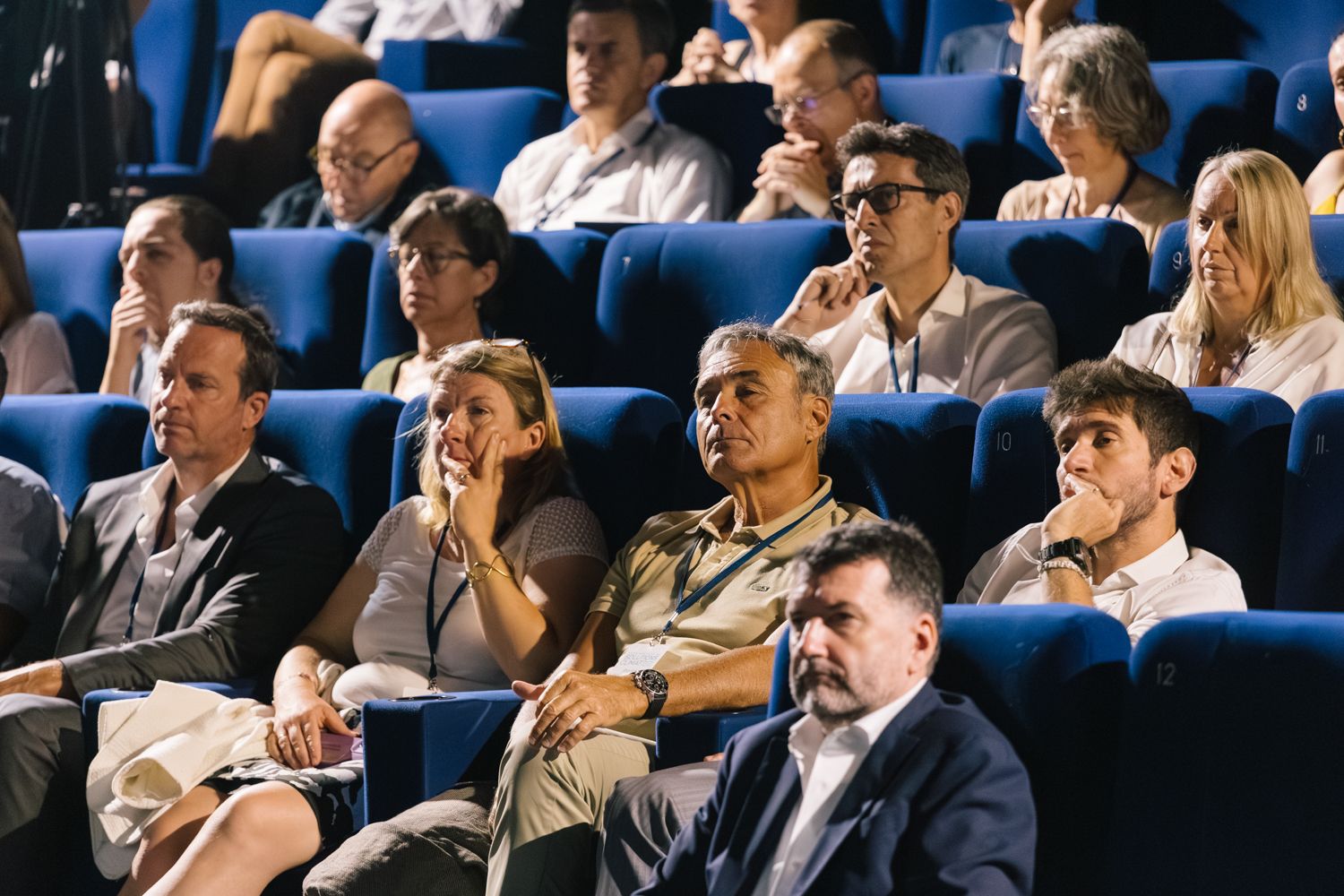 Audience members seated in blue chairs watch intently during an indoor event or presentation.