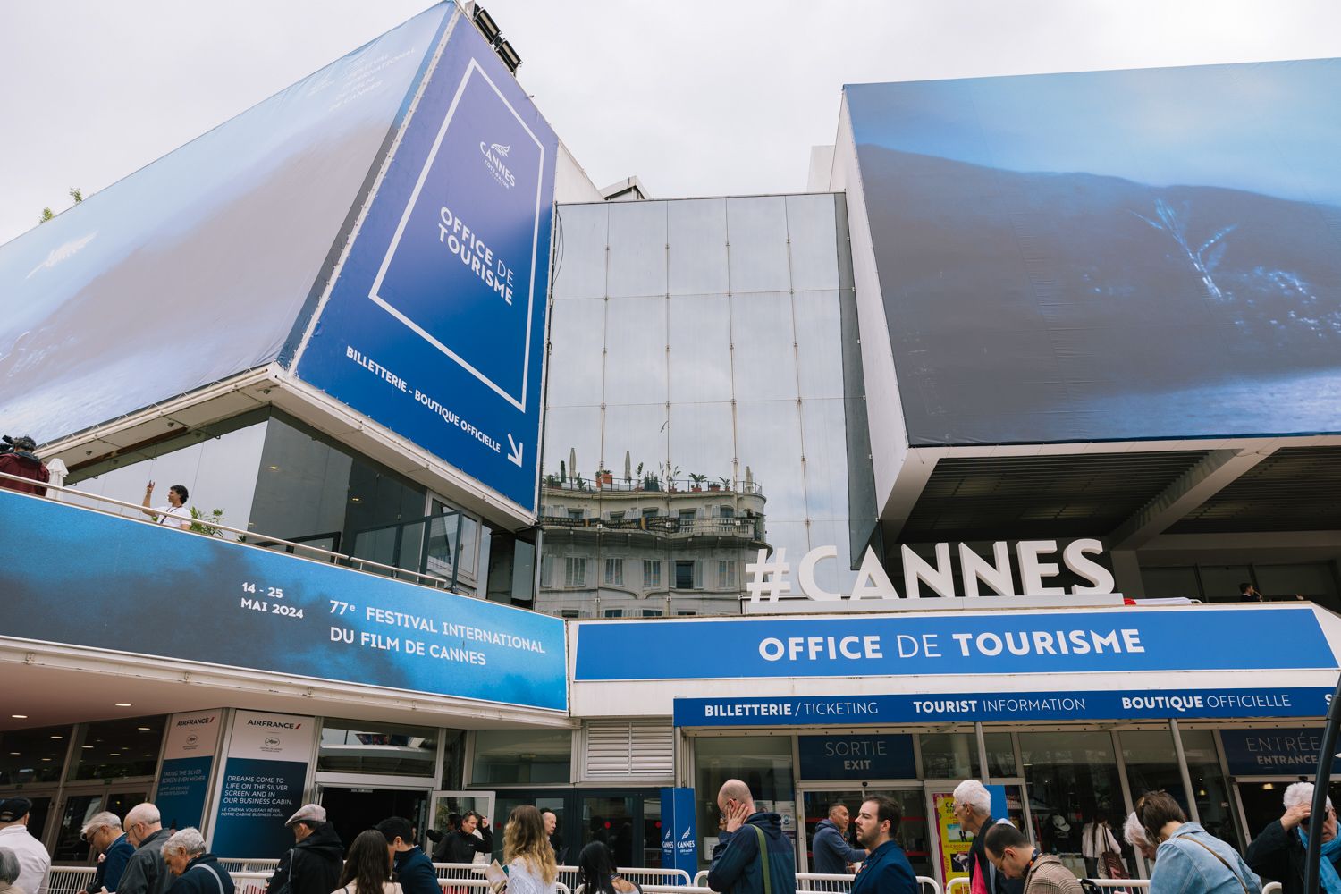 Exterior view of the Cannes Tourism Office with large promotional banners and crowd gathering.