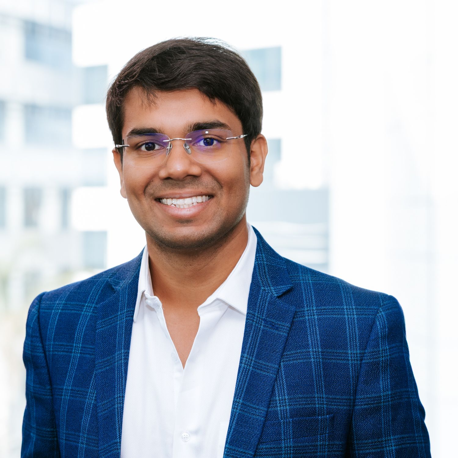Professional headshot of person wearing blue plaid blazer and white shirt against blurred office background.