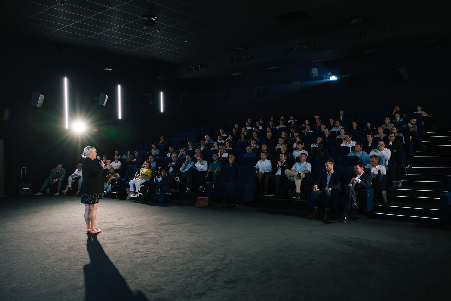 A presenter speaks to a packed audience in a darkened auditorium during a professional presentation.