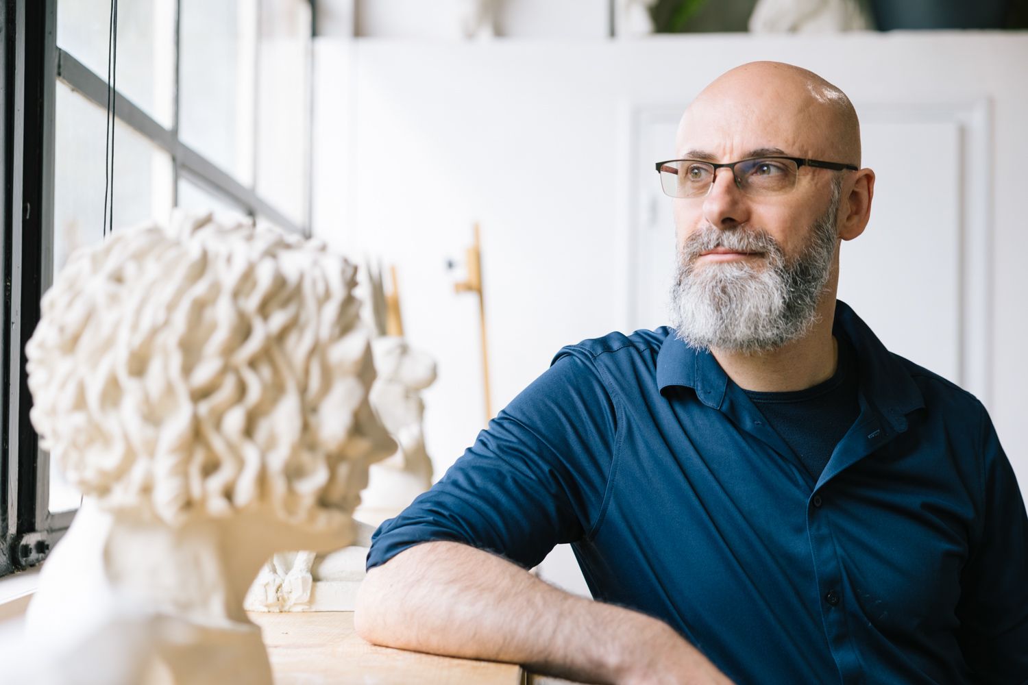 A bearded artist in glasses wearing a blue shirt sits contemplatively in a studio setting.