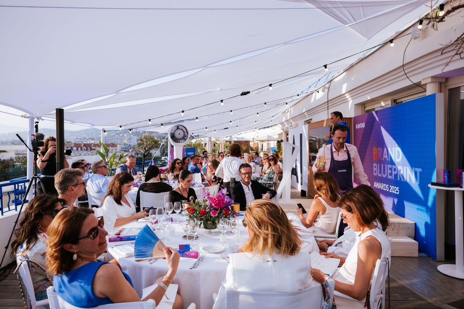 People seated at white tables during an outdoor networking event under string lights and white tent coverings.