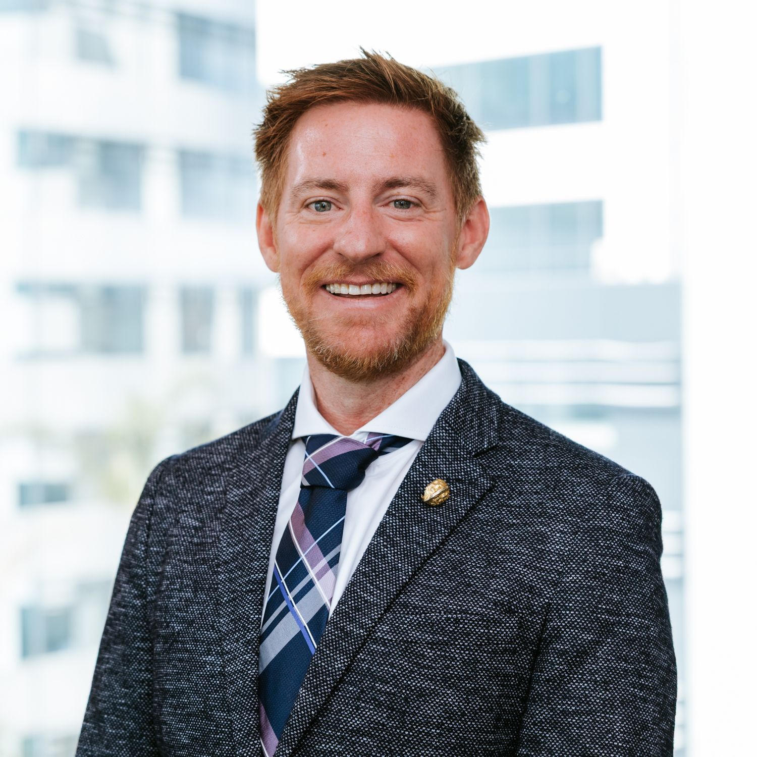 Professional wearing a gray suit and blue striped tie smiles warmly in a bright modern office setting.