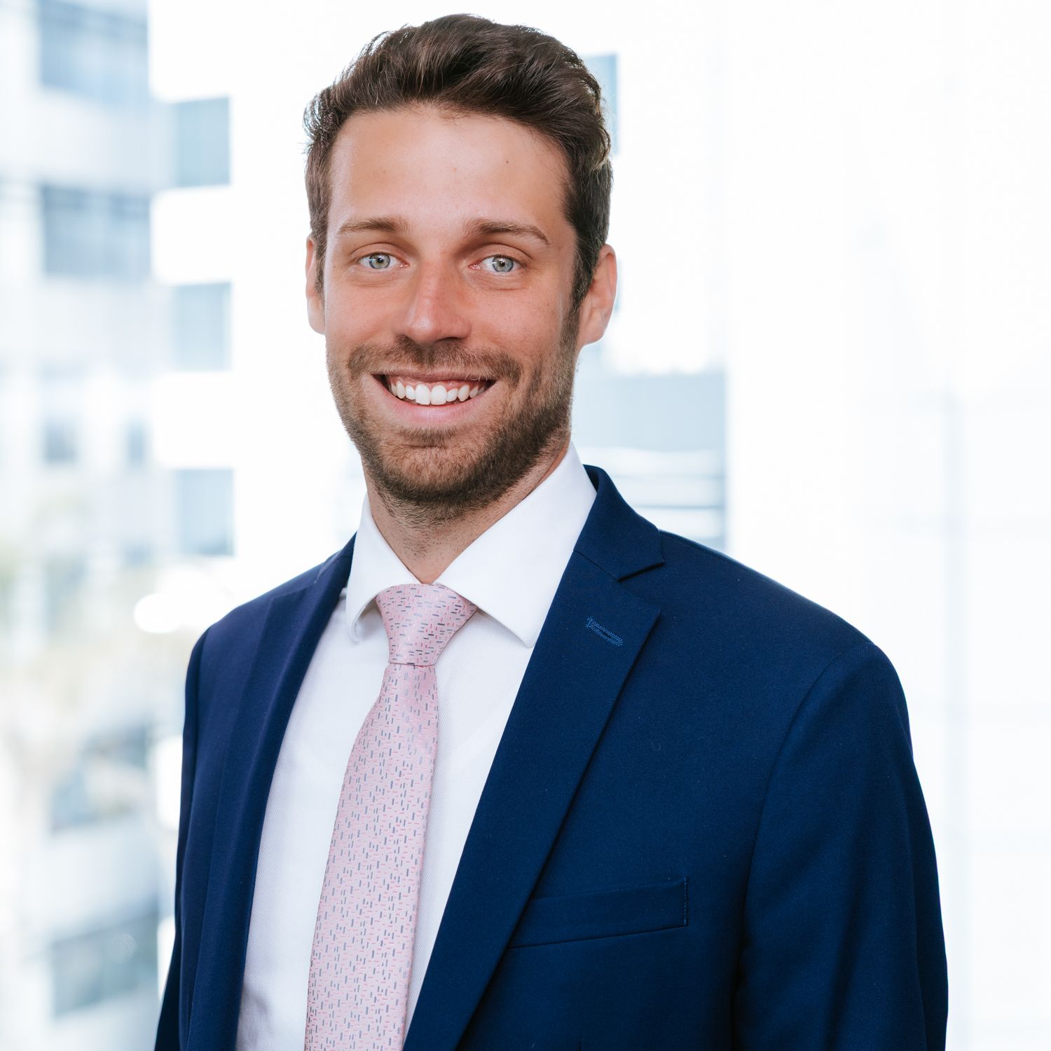 Professional wearing a navy suit and pink tie smiles warmly against a bright office background.