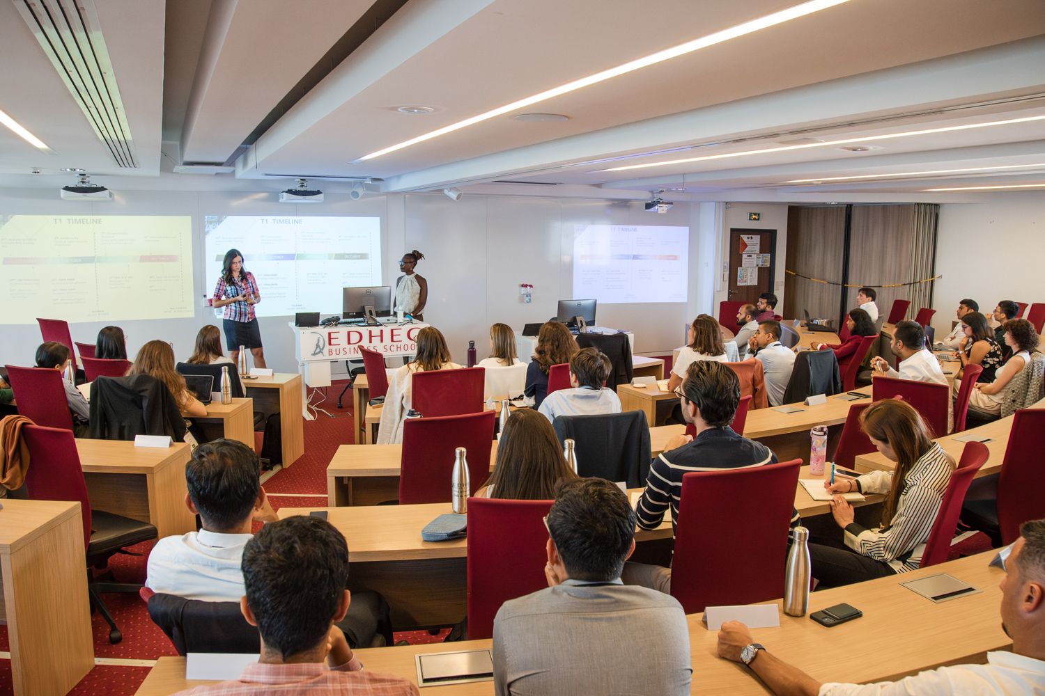 Modern lecture hall filled with students attending a presentation in red chairs.