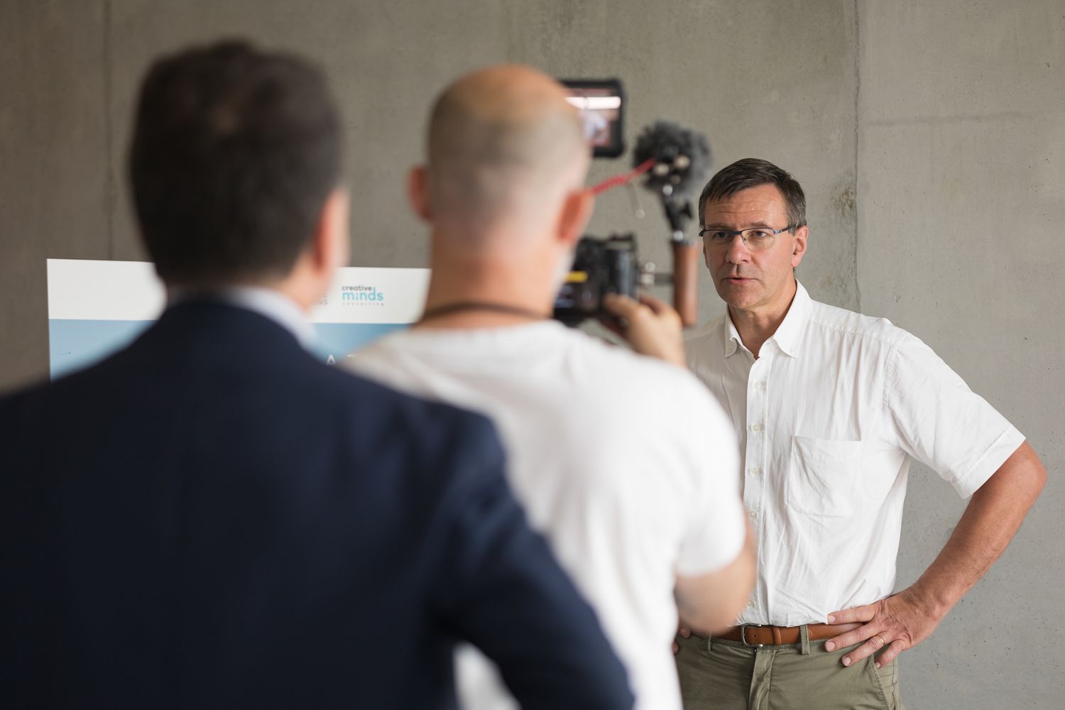 A filming session with camera crew and interviewee against a gray wall.