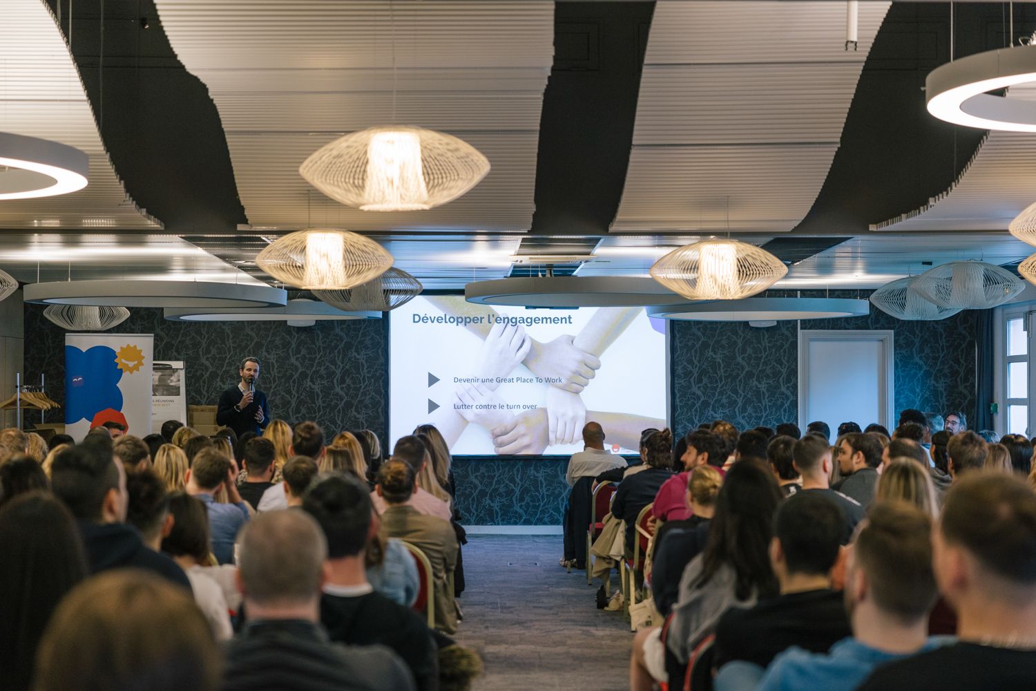 Large audience seated in a modern conference room with pendant lighting and projection screen.