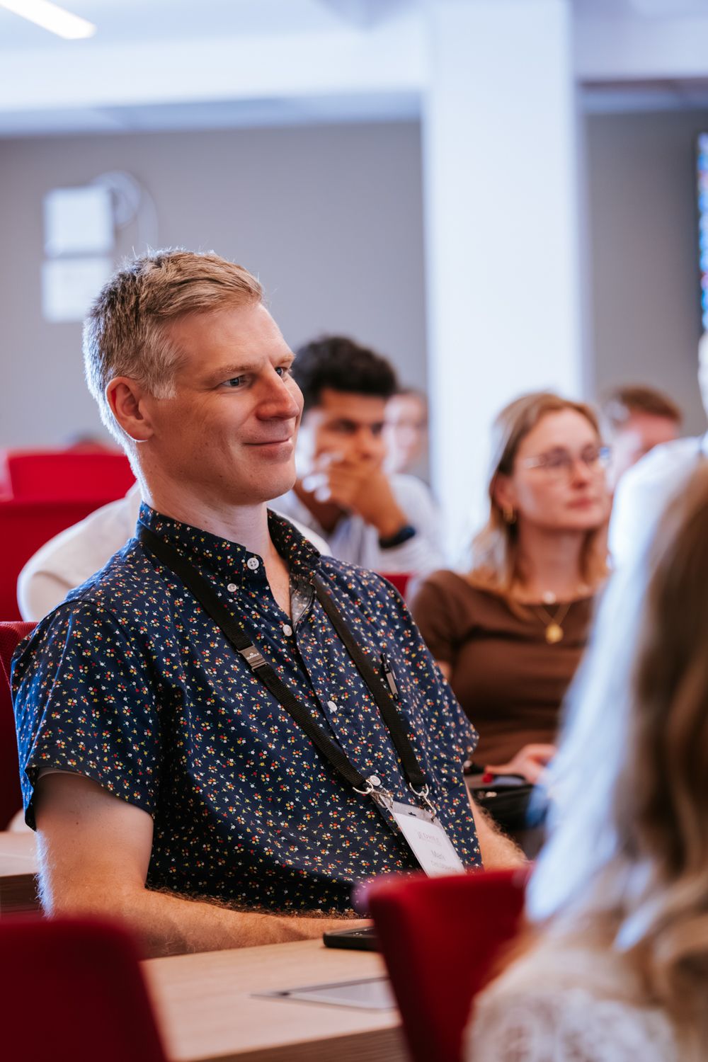 Person in polka dot shirt sitting in audience at conference or event with red chairs.