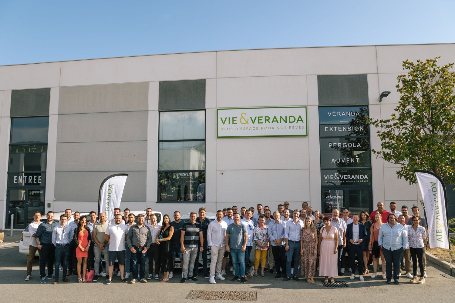 Large group photo of company employees standing outside corporate building with Vie & Veranda signage.