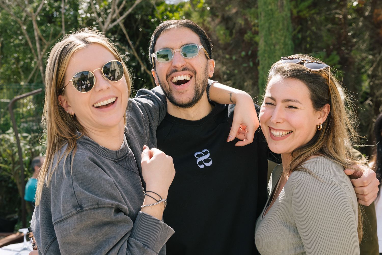 Three friends wearing sunglasses laughing together outdoors on a sunny day.