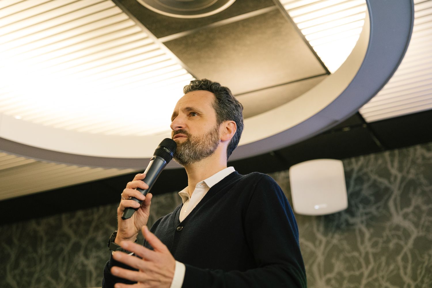 A speaker presents to an audience in a conference room with modern circular lighting fixtures.