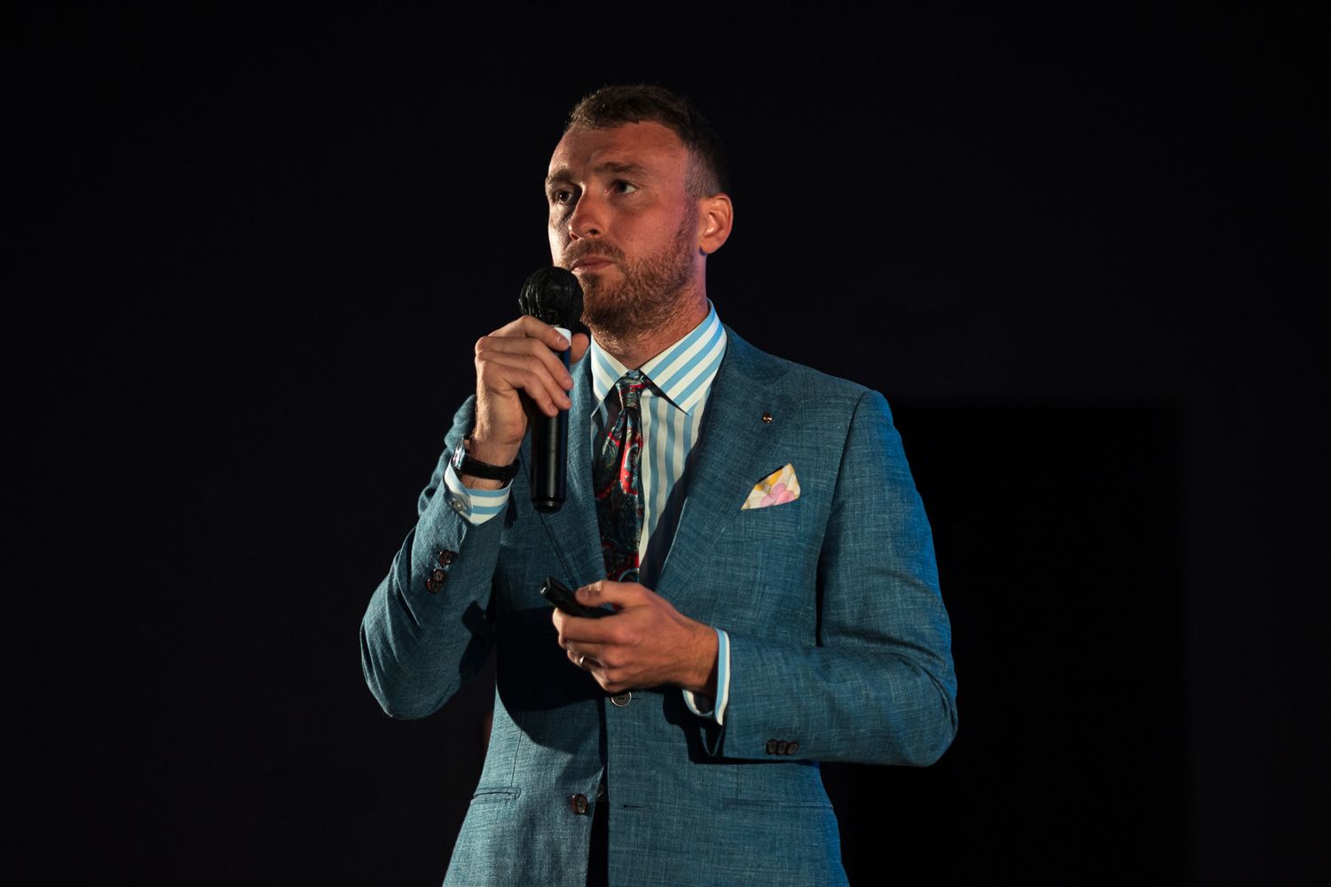 A speaker in a teal suit and tie presenting on stage against a dark background.