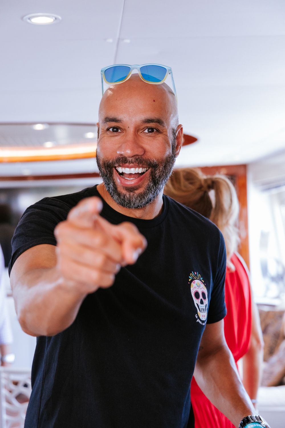 A person in a black t-shirt points and smiles at the camera in a bright indoor setting.