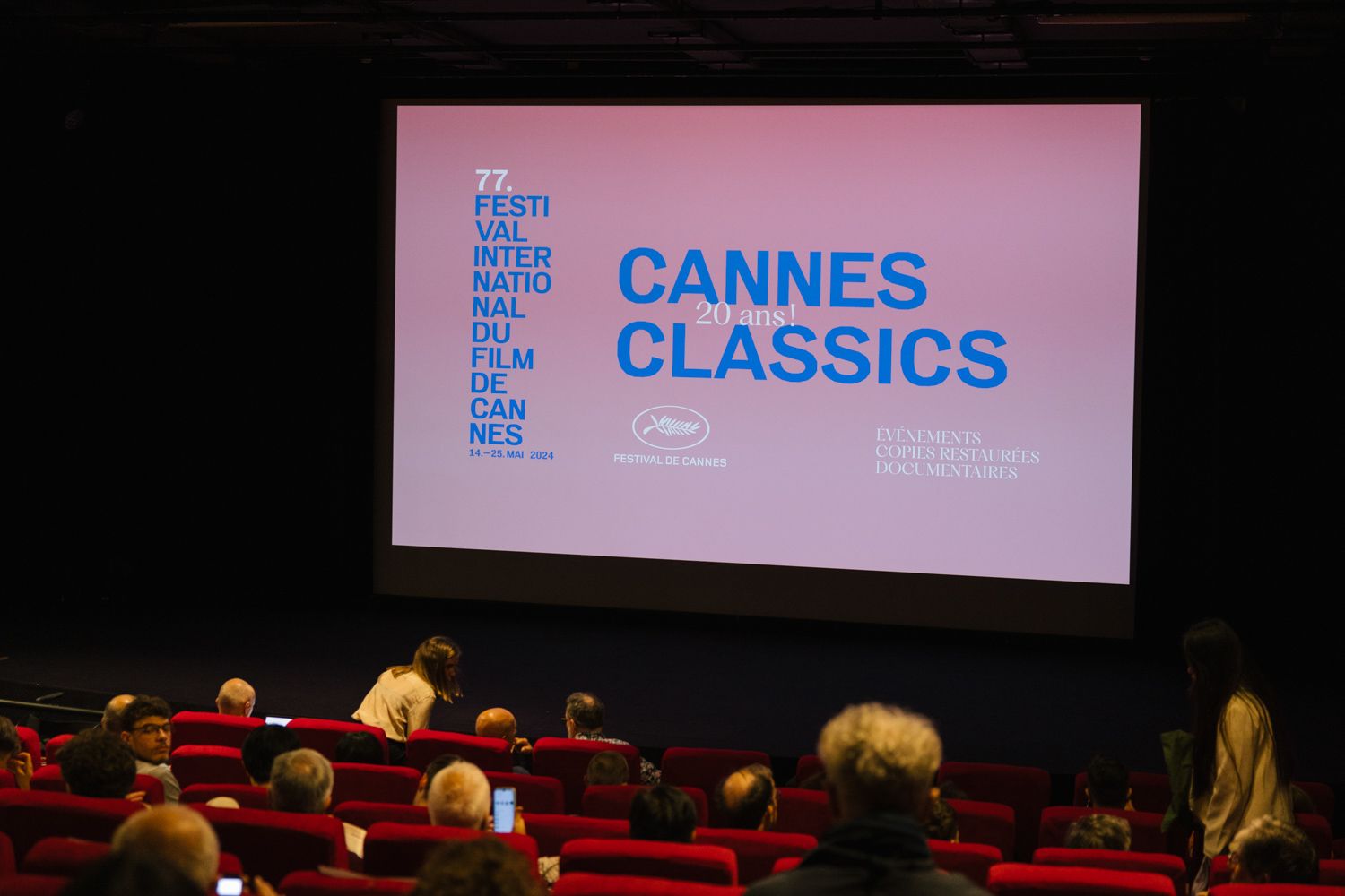 Movie theater audience watching a Cannes Classics screening with pink projection screen.