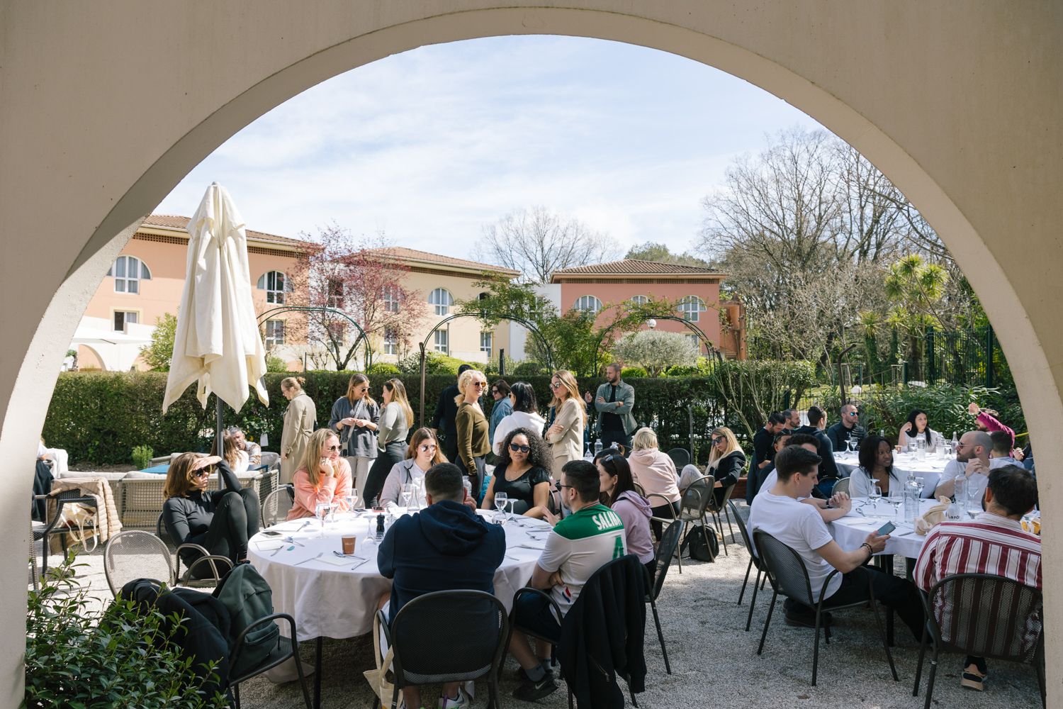 Outdoor dining scene under an arched colonnade with multiple tables of guests.