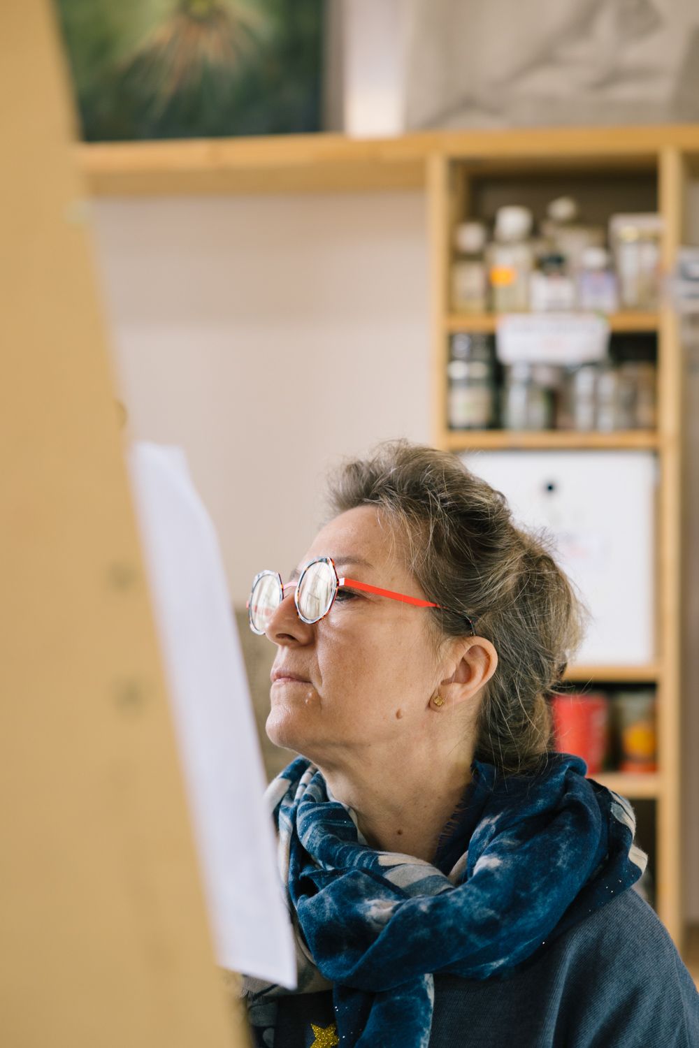 Artist in glasses and blue scarf looking up while working in studio.