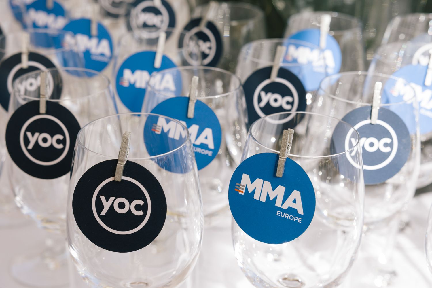 Wine glasses decorated with MMA and YOC branded stickers arranged for an event.