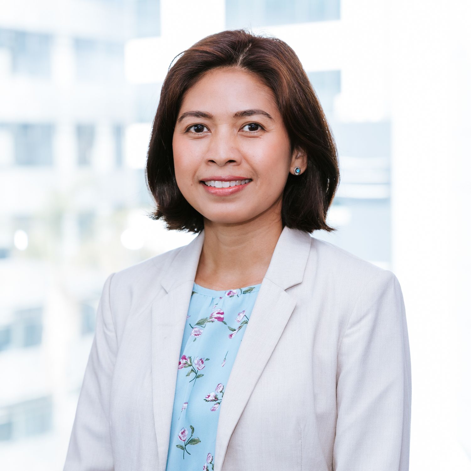 Professional woman in light gray blazer and blue floral top smiling against bright office background.
