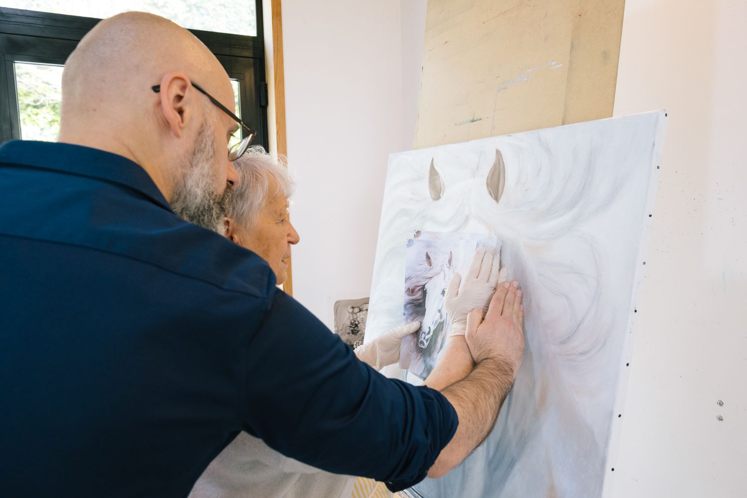 An artist in a navy shirt works on a detailed sketch pinned to a wall in a bright studio space.