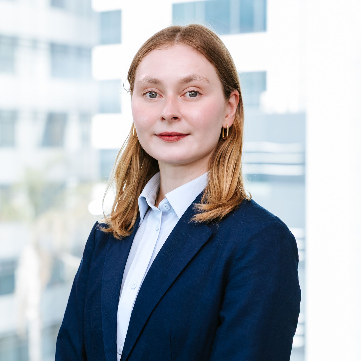 Professional portrait of a person in a navy blazer and white button-down shirt against a bright office background.