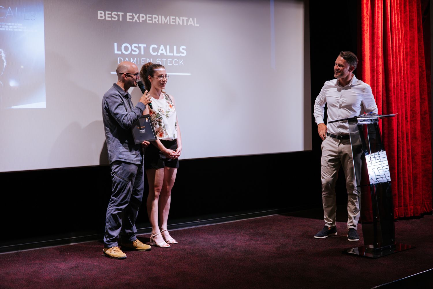 People stand on stage at an awards ceremony for Best Experimental category with red curtain and projection screen visible.