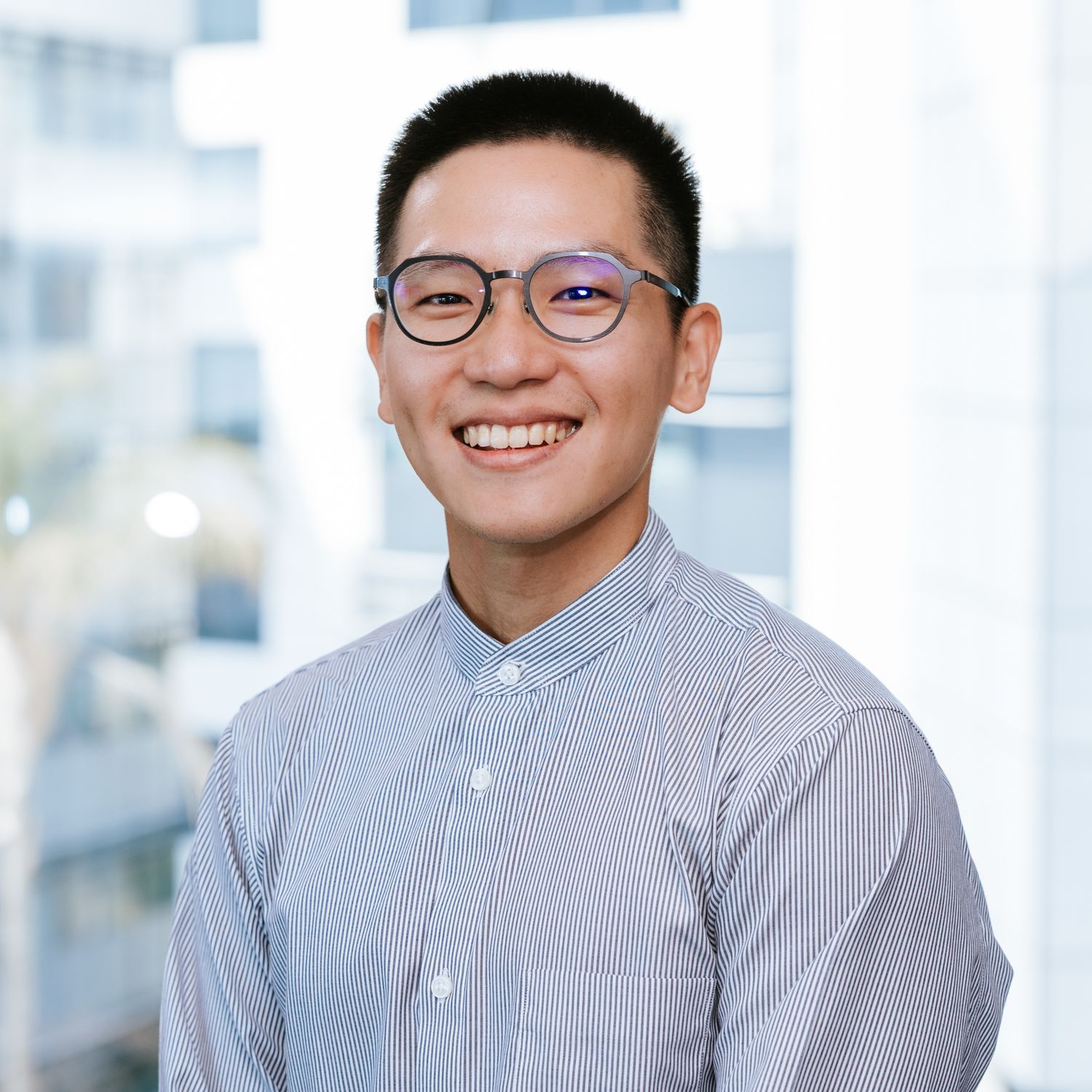 Professional headshot portrait of someone wearing glasses and a striped button-up shirt in an office setting.