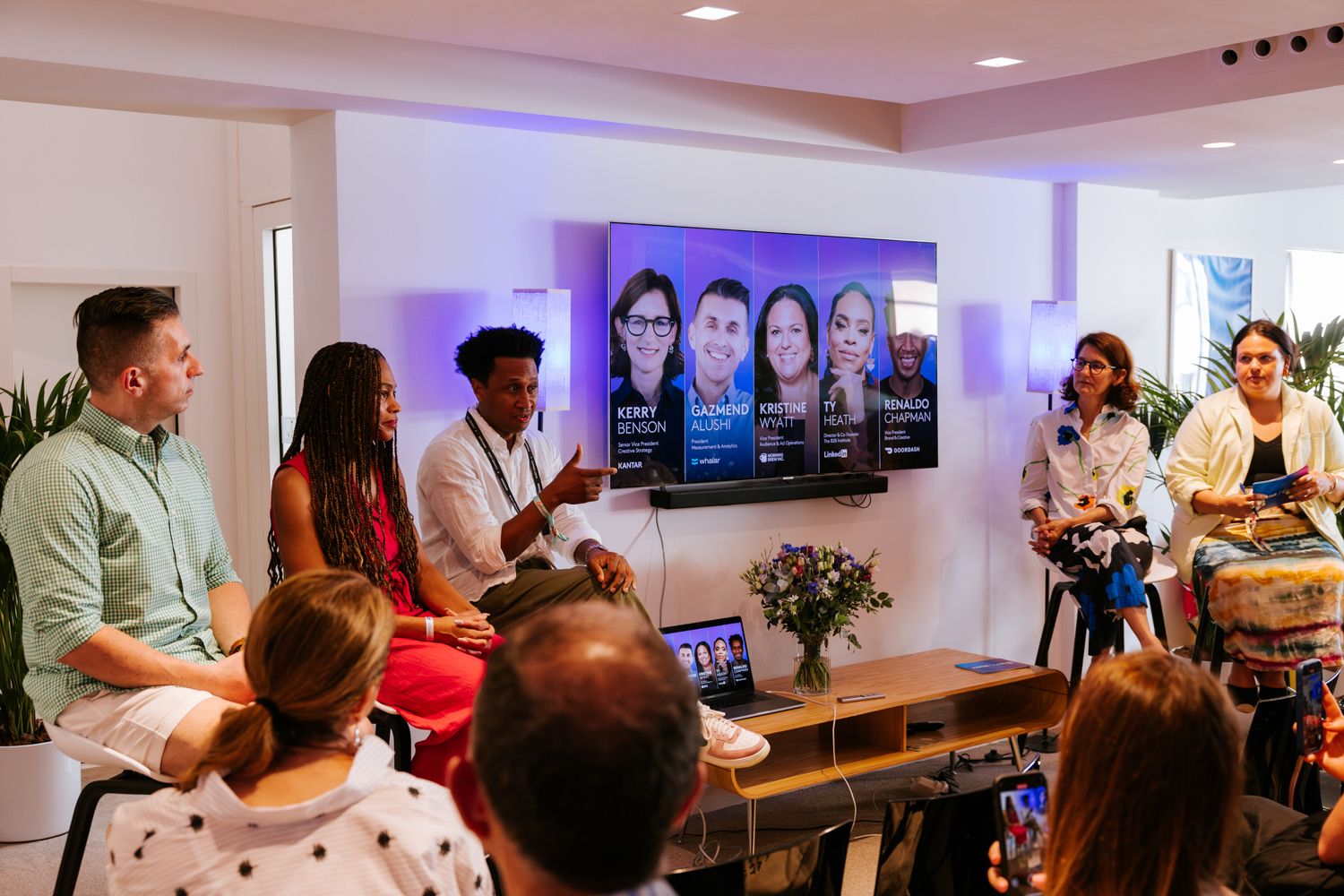 A group gathers in a modern office space for a presentation with a digital display showing profiles on the wall.