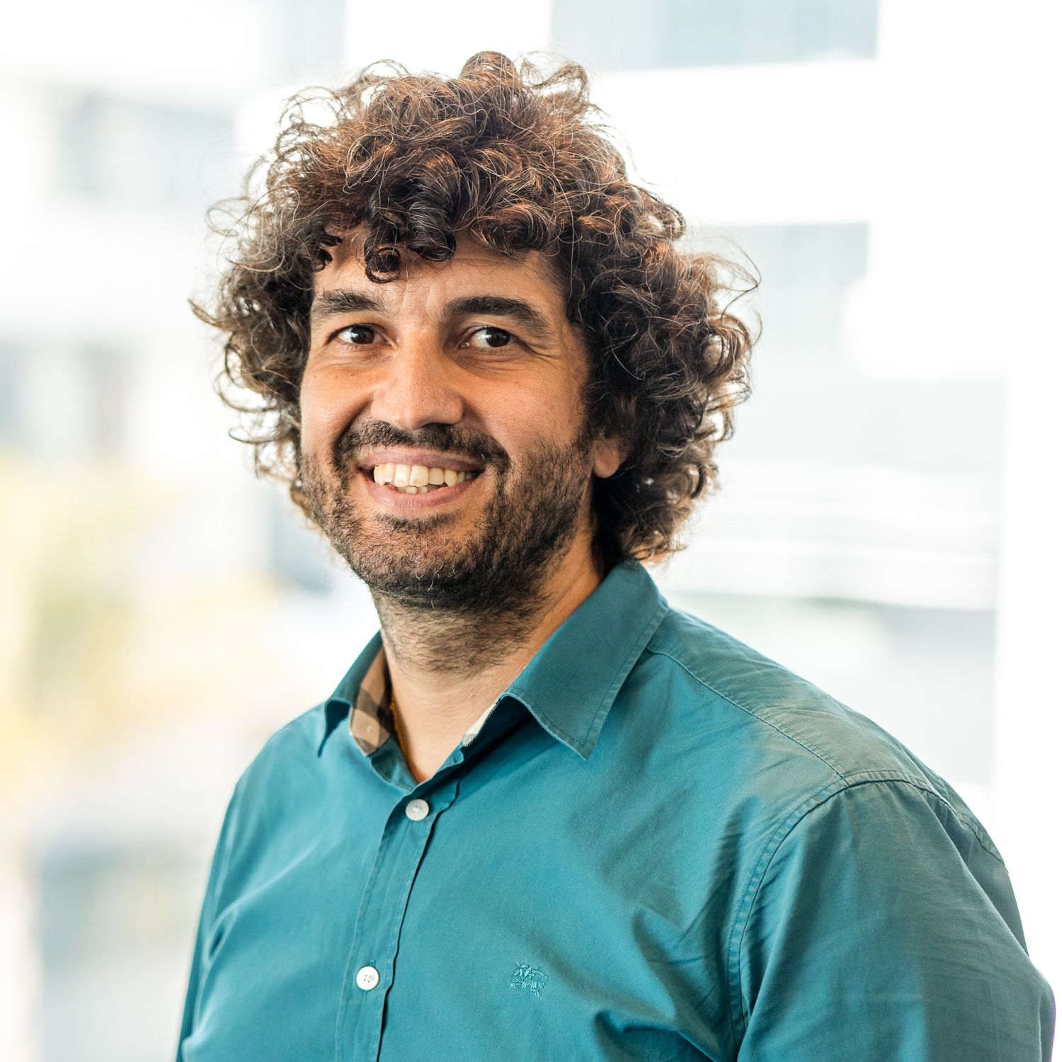 Professional headshot of a person in teal shirt with curly hair against office background.