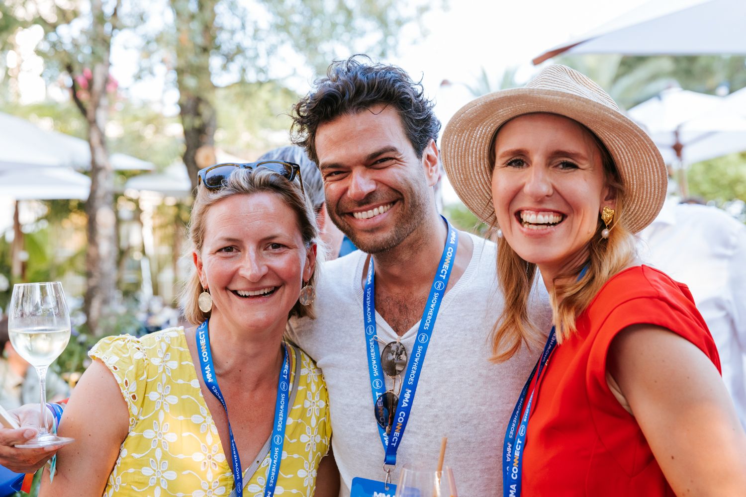 Three smiling people wearing event lanyards and summer attire enjoying an outdoor gathering.