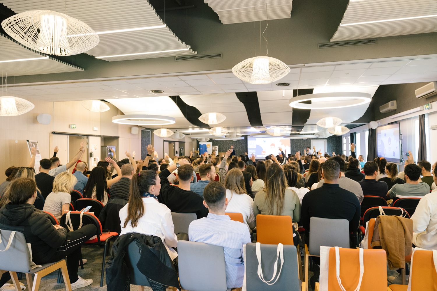 A large audience sits in rows of chairs during a conference presentation in a modern meeting space.