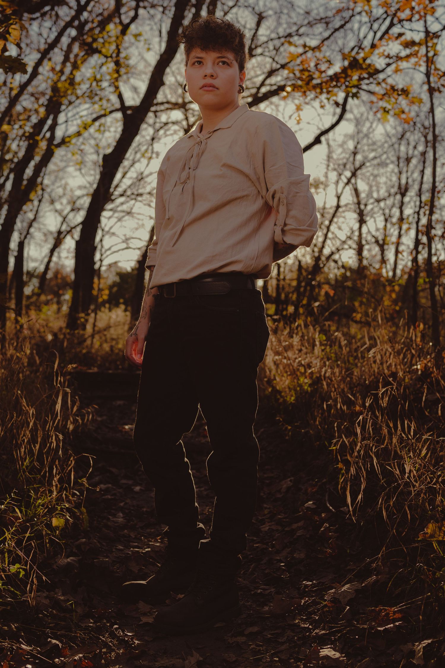 A person in a beige shirt and dark pants stands on a forest trail during golden hour.
