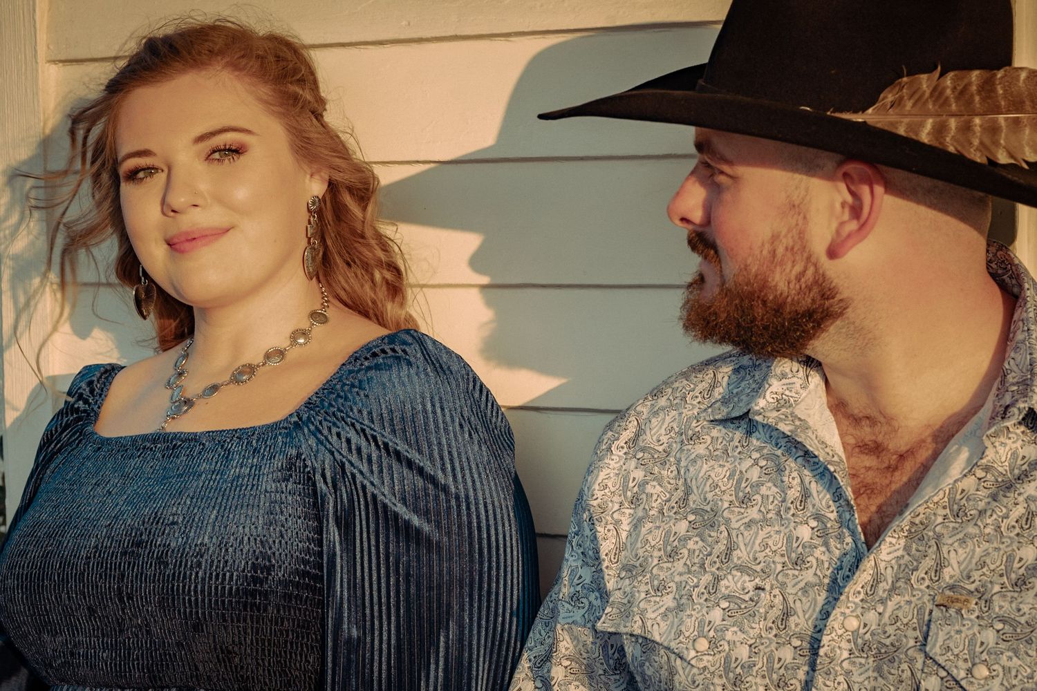 A country couple in western attire posing together against a light blue wall.