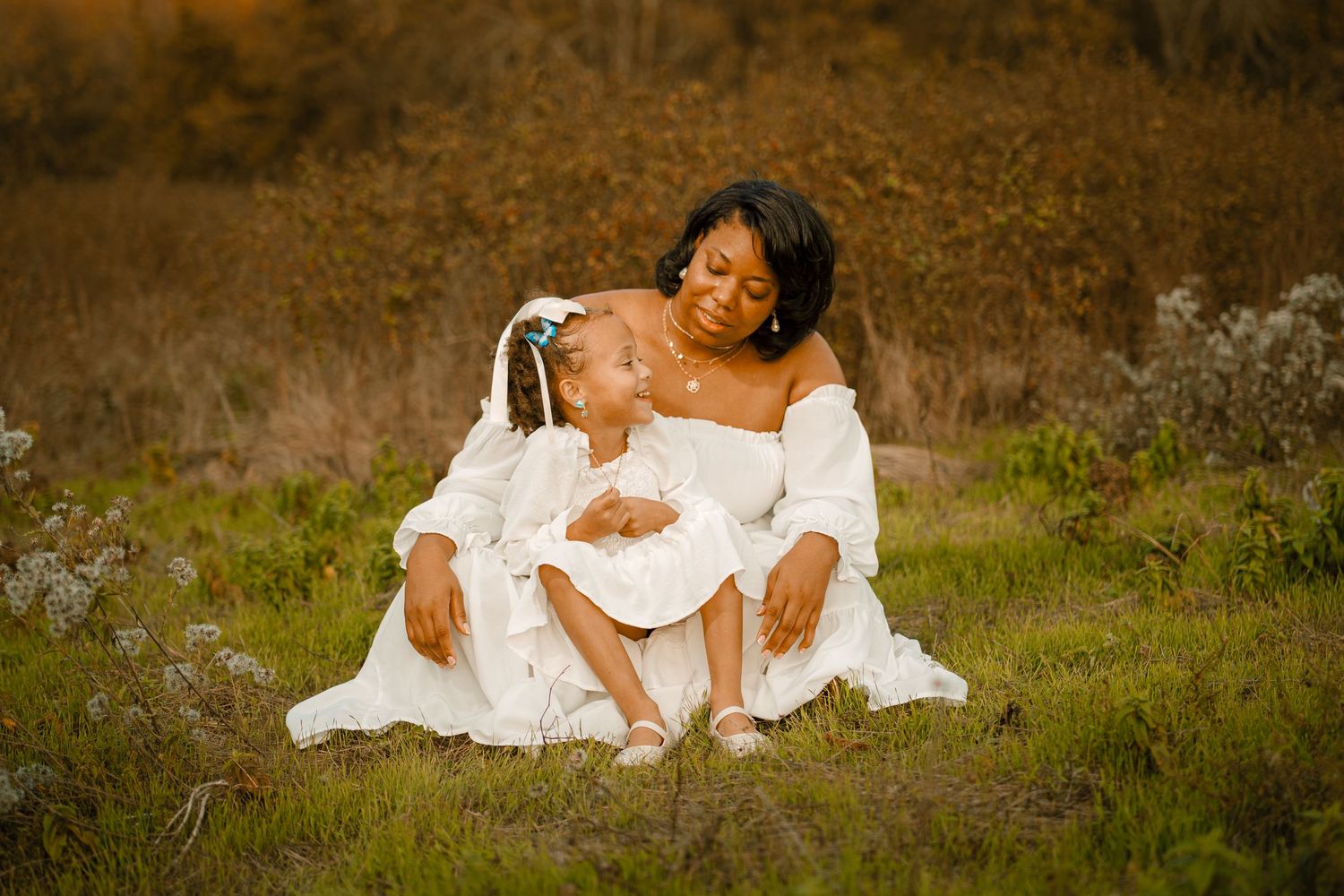 Two people in white dresses sit together on grass during golden hour in a romantic outdoor photoshoot.