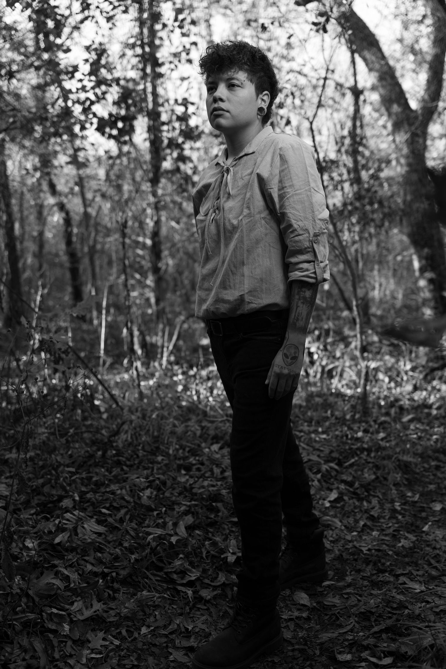 A black and white portrait showing a person in vintage attire standing in a woodland setting.