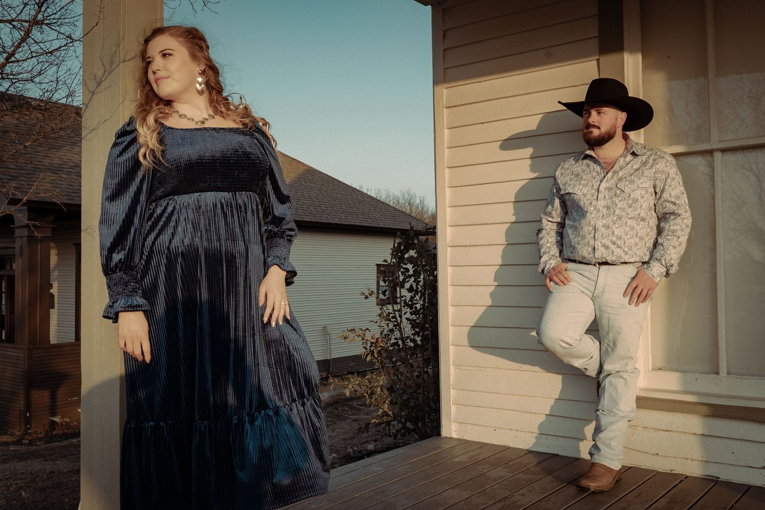 A couple in western attire pose separately against a house exterior in natural light.
