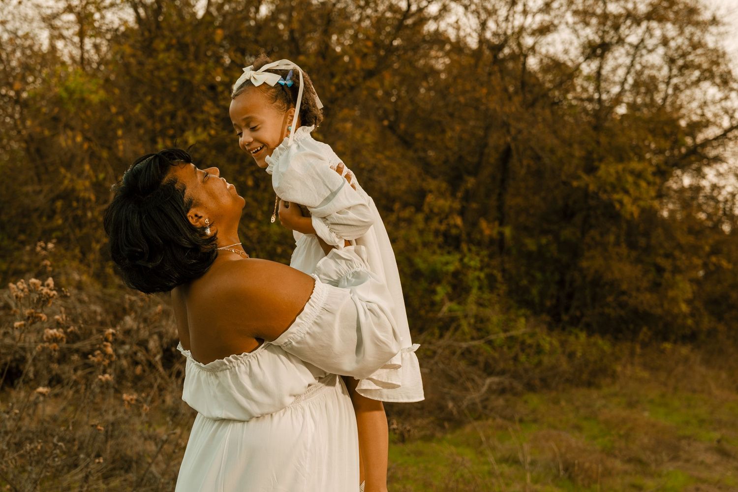 Mother in white dress lifting child during golden hour photoshoot.