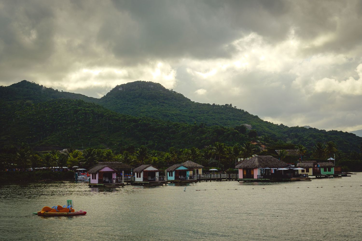 Stilt houses line a waterfront with mountains and dramatic cloudy sky in the background.