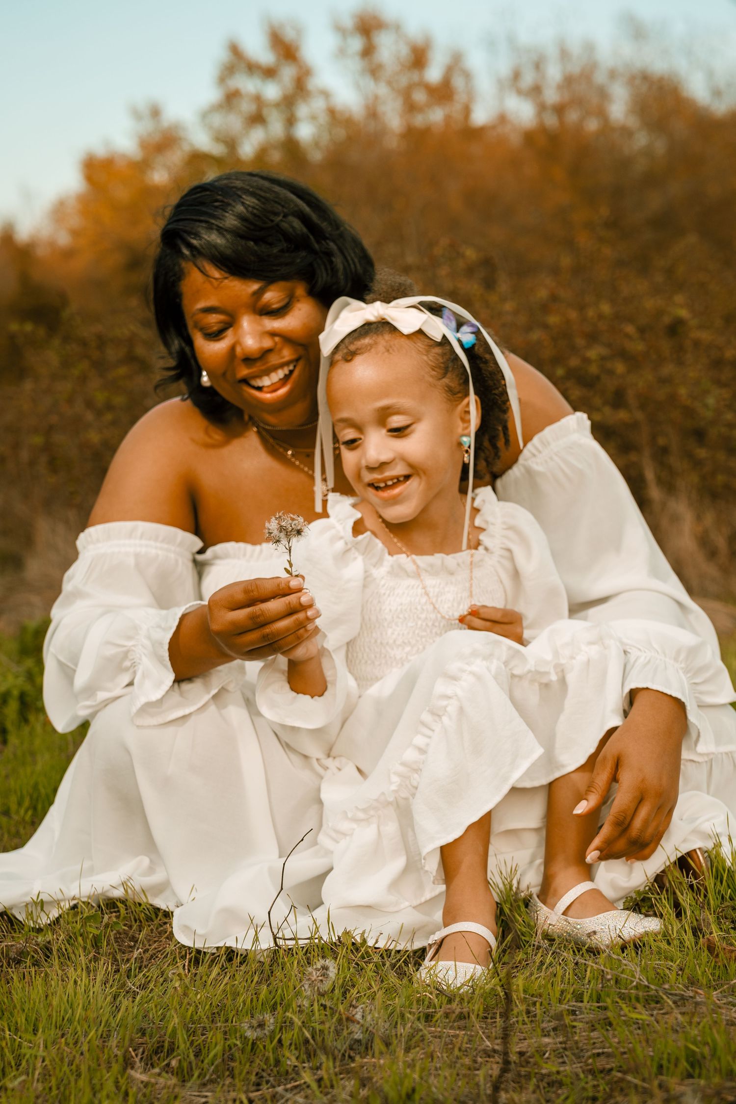 Two figures in flowing white dresses share a joyful moment together while sitting in a sunlit field at golden hour.