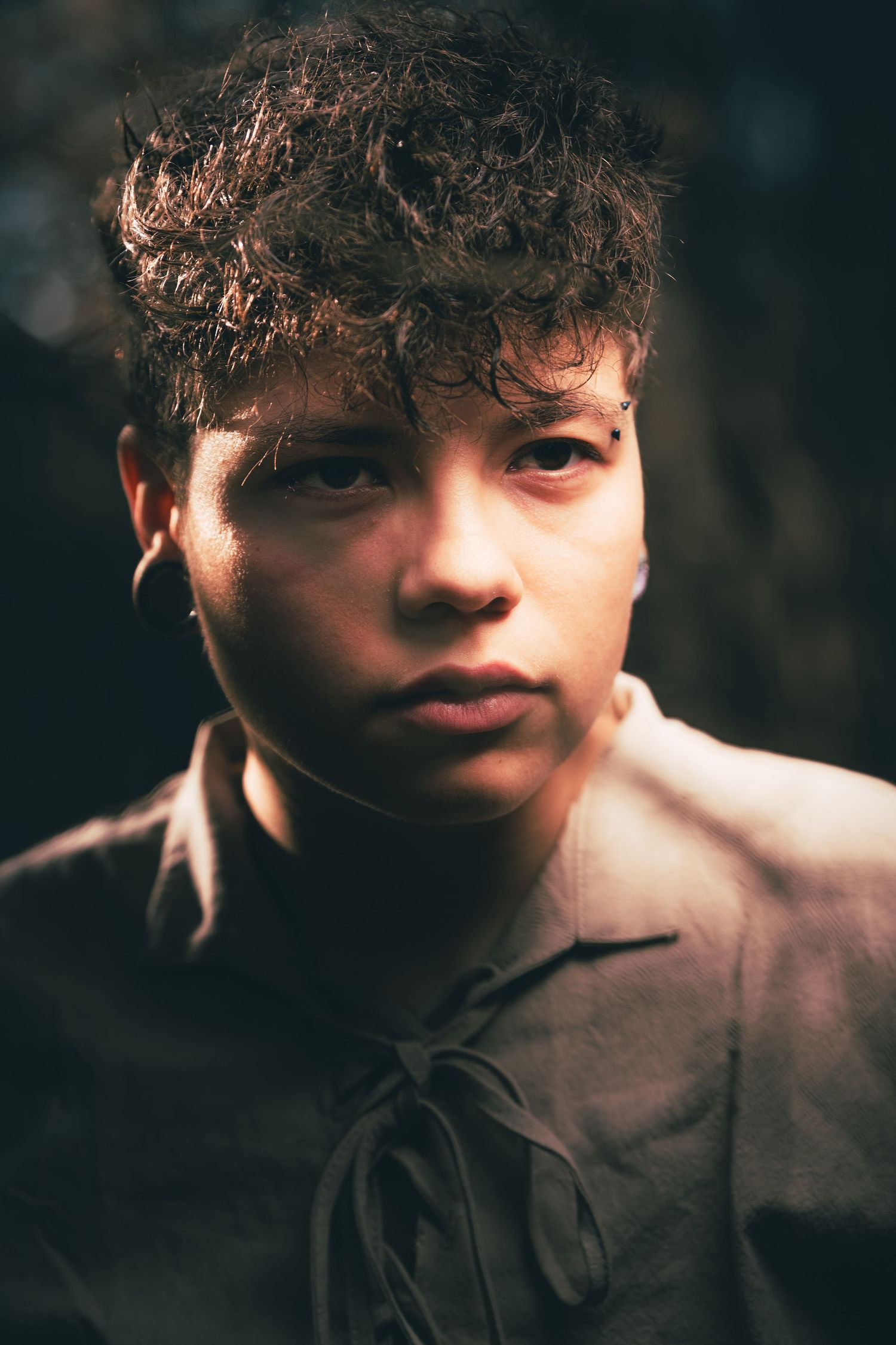 Artistic portrait in moody lighting showing curly hair and tan shirt against dark background.