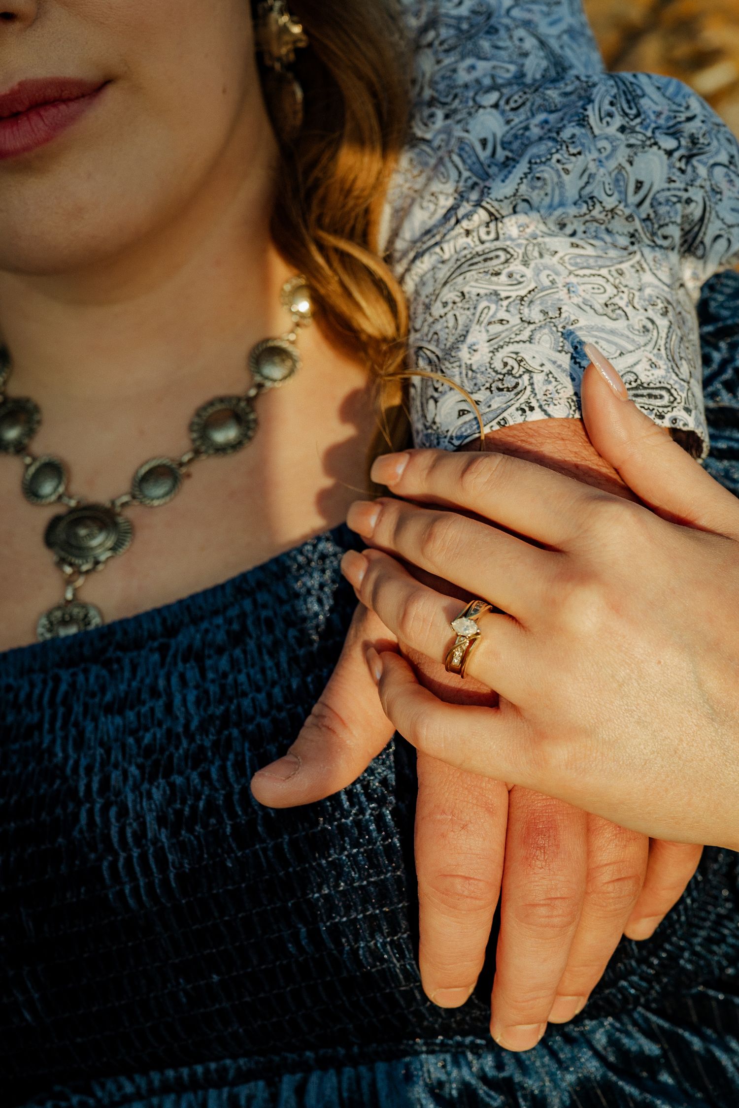 Close-up of hands wearing an engagement ring with a teal dress and statement necklace in view.
