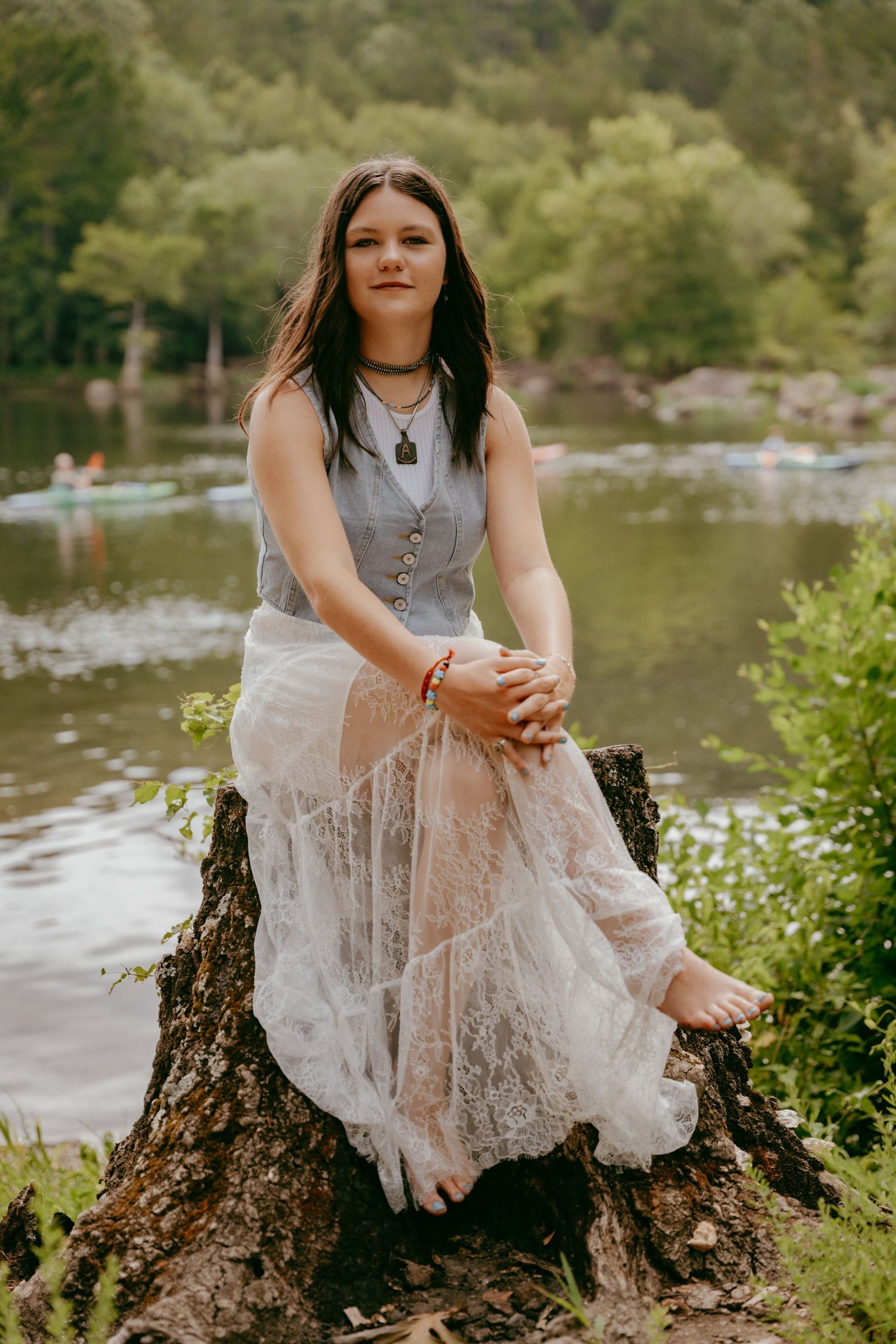 A person wearing a denim vest and flowing white lace skirt sits on a tree stump by a serene lake in nature.